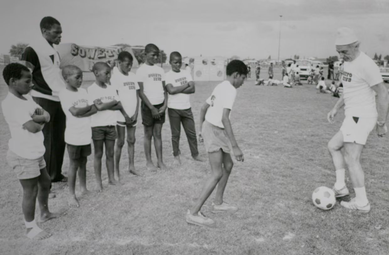 Stanley Matthews dribbles a football while a line of boys in Soweto club shirts watch
