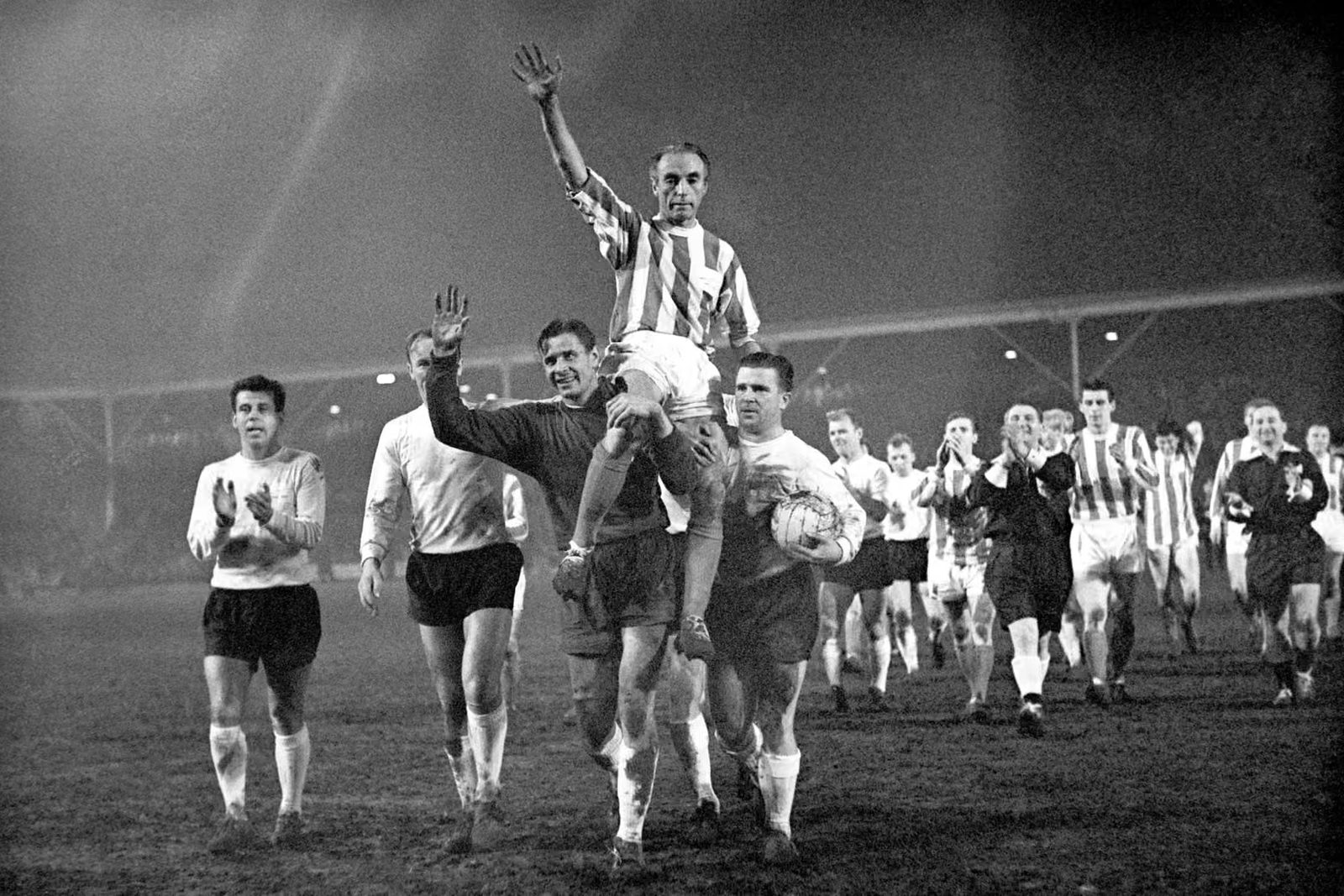 Sir Stanley Matthews is chaired off the pitch, arms raised, as players applaud in the mist